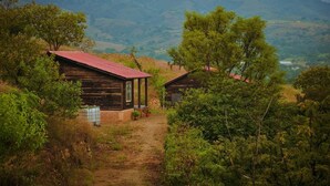 Una cabaña rústica con techo rojo, rodeada de abundante vegetación y colinas al fondo.