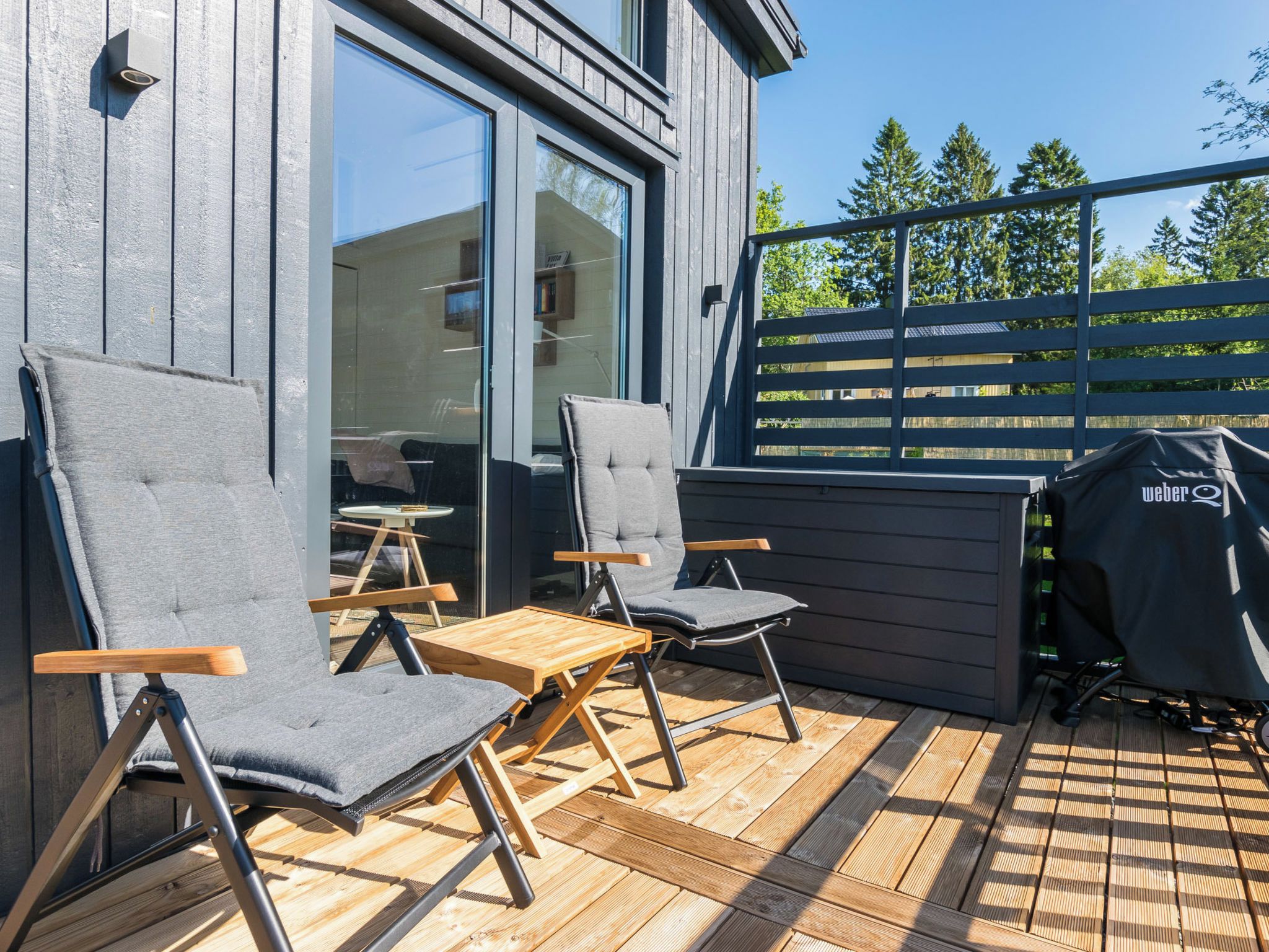 Un espace extérieur aménagé avec deux chaises et une table, un barbecue, et une vue sur une terrasse en bois et des arbres.