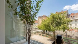 A window with a vase of branches, a lamp, and a view of buildings and a river. - Tromostovje III at Triple Bridge Ljubljana, Ljubljana, Slovenia (Ljubljana)