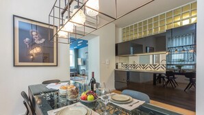 Uma área de refeições moderna com uma mesa de vidro, cadeiras de metal e um candelabro. Uma cozinha pode ser vista ao fundo, com uma janela grande.