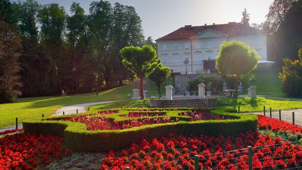 Um jardim formal com sebes aparadas, flores vermelhas e um edifício clássico ao fundo.