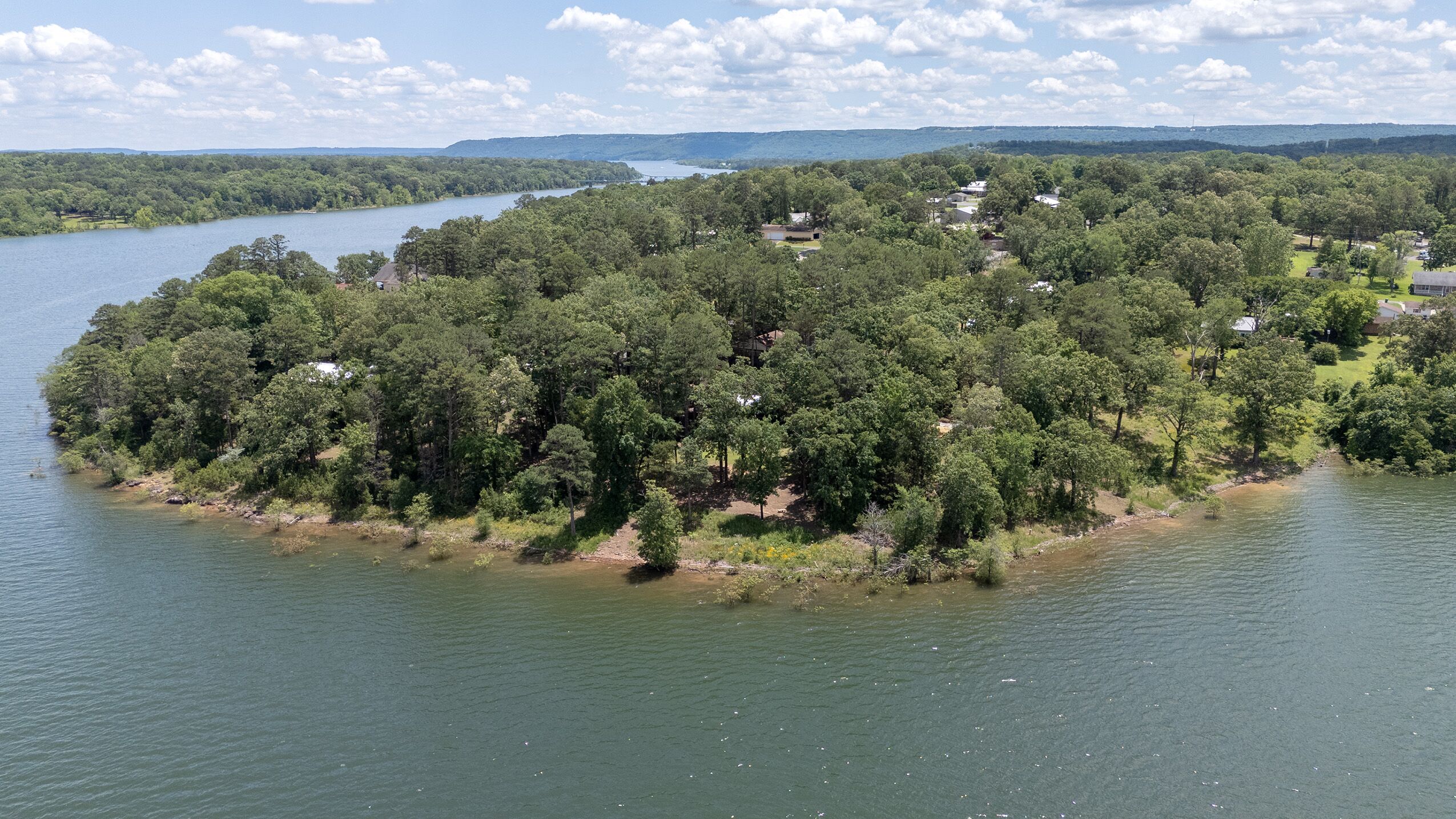 Une vue aérienne d’une île boisée avec un lac, entourée d’une zone résidentielle avec des maisons, et une vue lointaine sur une étendue d’eau et des collines.