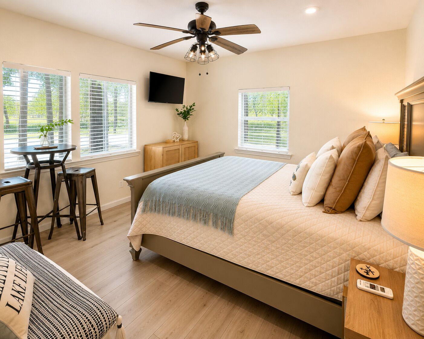 A bedroom with a bed, a desk, two chairs, a TV, and a ceiling fan.
