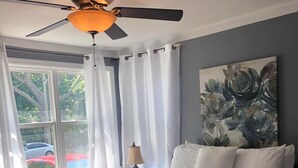 Een slaapkamer met een bed, een plafondventilator, een raam met gordijnen en een schilderij aan de muur.