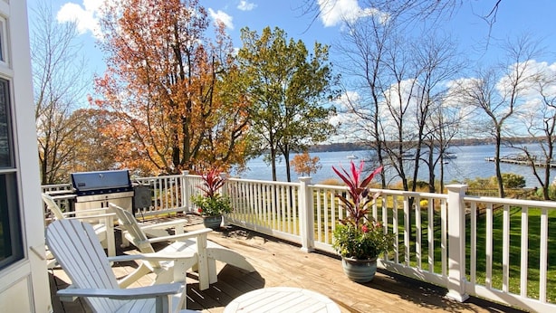 A wooden deck with white chairs and a table, overlooking a lake with autumn foliage.