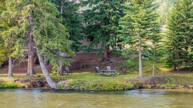 A lakeside view with a picnic table and chairs, surrounded by trees and a building in the background. - Clarks Fork Crossing- Direct River Access, Large family Cabin! (Cody)