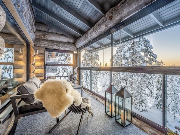 A cozy interior with a wooden ceiling, a sofa, a fluffy white object, and a view of snow-covered trees.