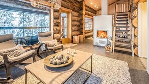 L’intérieur d’une cabane en rondins, avec une cheminée, un coin salon et une vue sur des arbres couverts de neige.