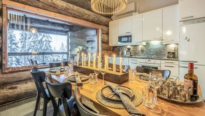 Une cuisine moderne avec des meubles blancs, une table à manger en bois agrémentée de bougies, et une vue sur les arbres enneigés à travers de grandes fenêtres.