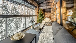 L’intérieur chaleureux d’un chalet avec une cheminée en pierre, un sapin de Noël et une grande fenêtre donnant sur une forêt enneigée.