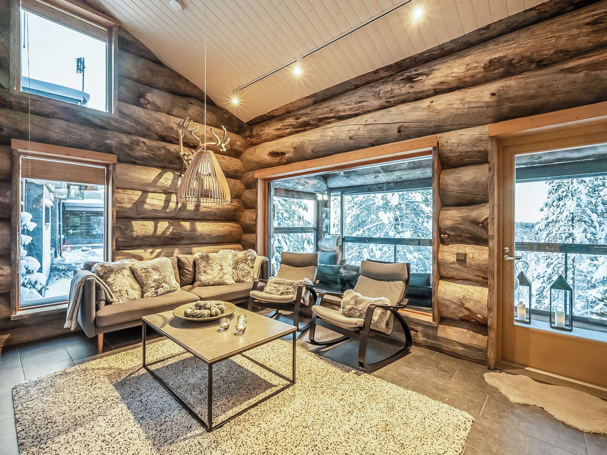 A log cabin interior with a sofa set, chairs, a coffee table, and large windows with snowy views.