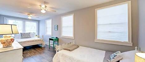 A bedroom with two beds, large windows, a ceiling fan, and a bedside table with a lamp.