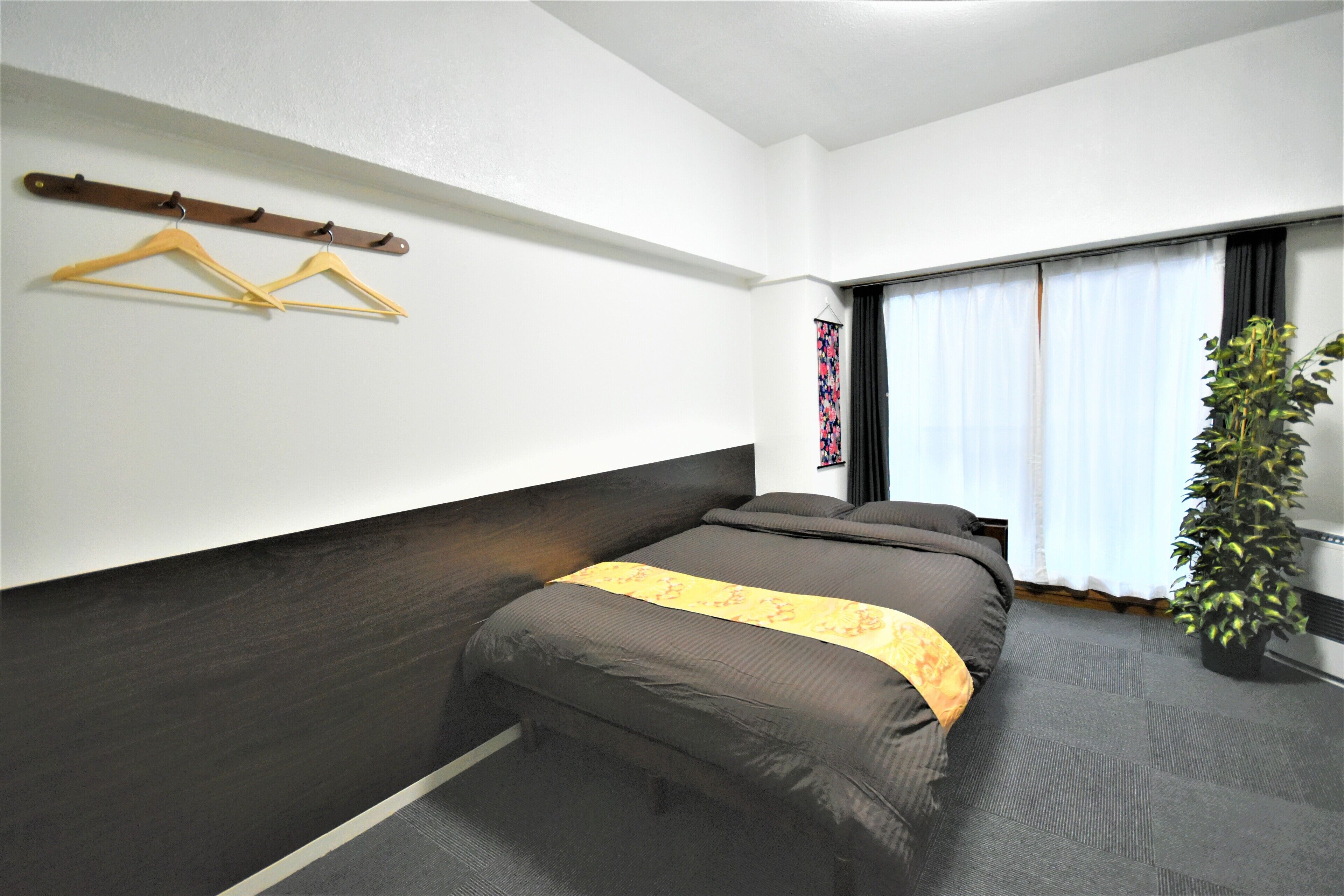 A bedroom with a bed, a coat rack with hangers, a window with curtains, and a potted plant.