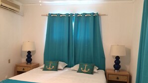 A neatly made bed with a white and teal bedspread, teal curtains, and matching pillows with anchor designs. There are two bedside tables with lamps and a small drawer. - Cute beach apartment (Savaneta)