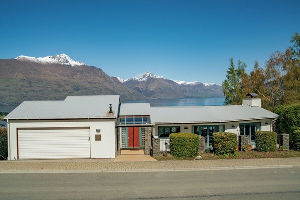 Une maison à deux étages avec un garage, une porte d’entrée rouge et une vue sur des montagnes et un lac en arrière-plan.