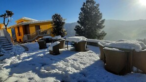 A snow-covered outdoor area with a yellow building, wicker furniture, and a view of distant hills. - juSTa Mukteshwar Retreat & Spa (Dhari)