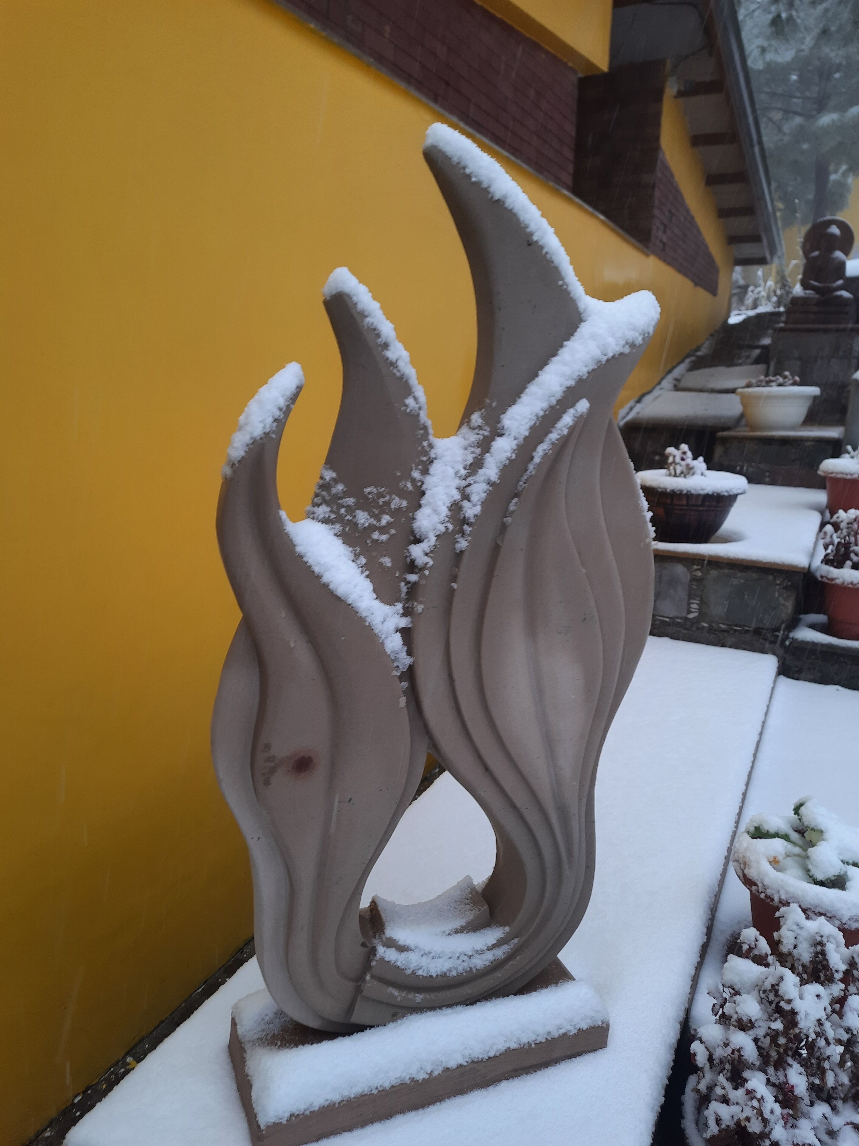 Una escultura cubierta de nieve en un patio con una pared amarilla y plantas en macetas.