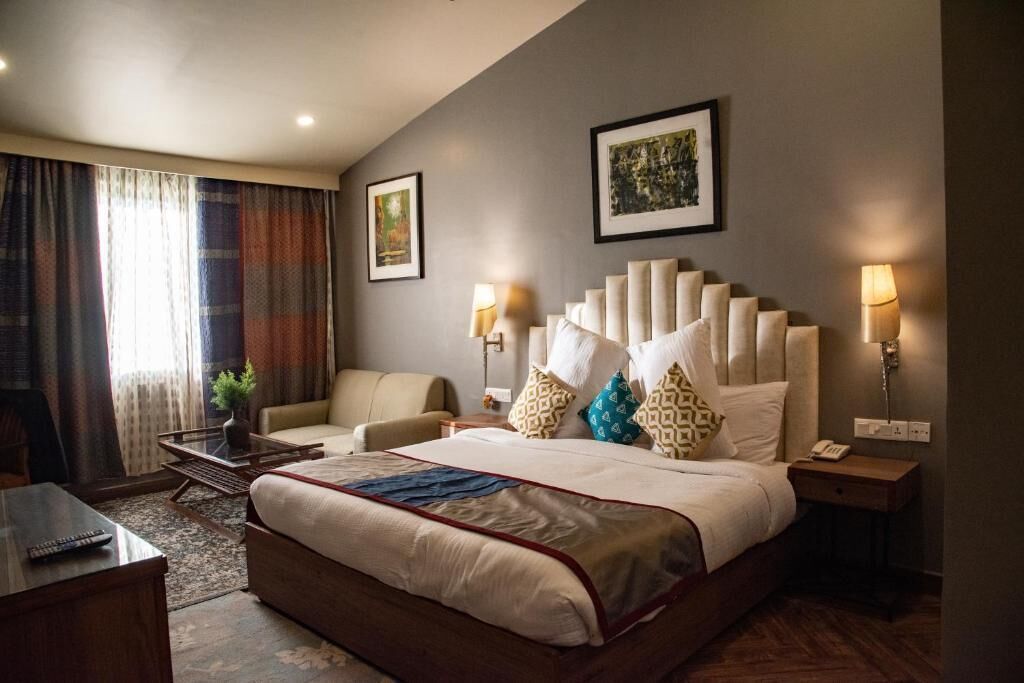 A hotel room with a large bed, a sofa, a desk, and a chair. There are two bedside tables with lamps and a small table with a plant.