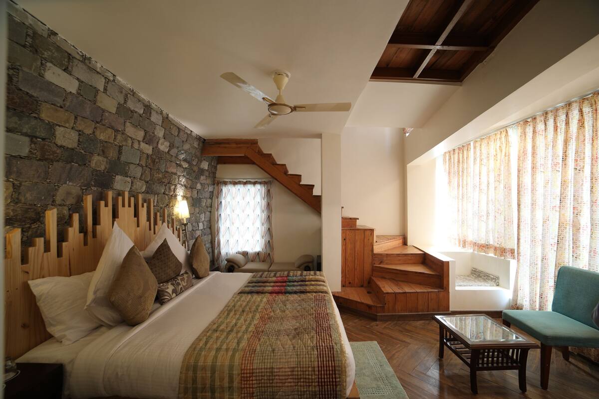 A bedroom with a stone wall, wooden headboard, a bed with multiple pillows, a wooden staircase, a glass table, and a green armchair.