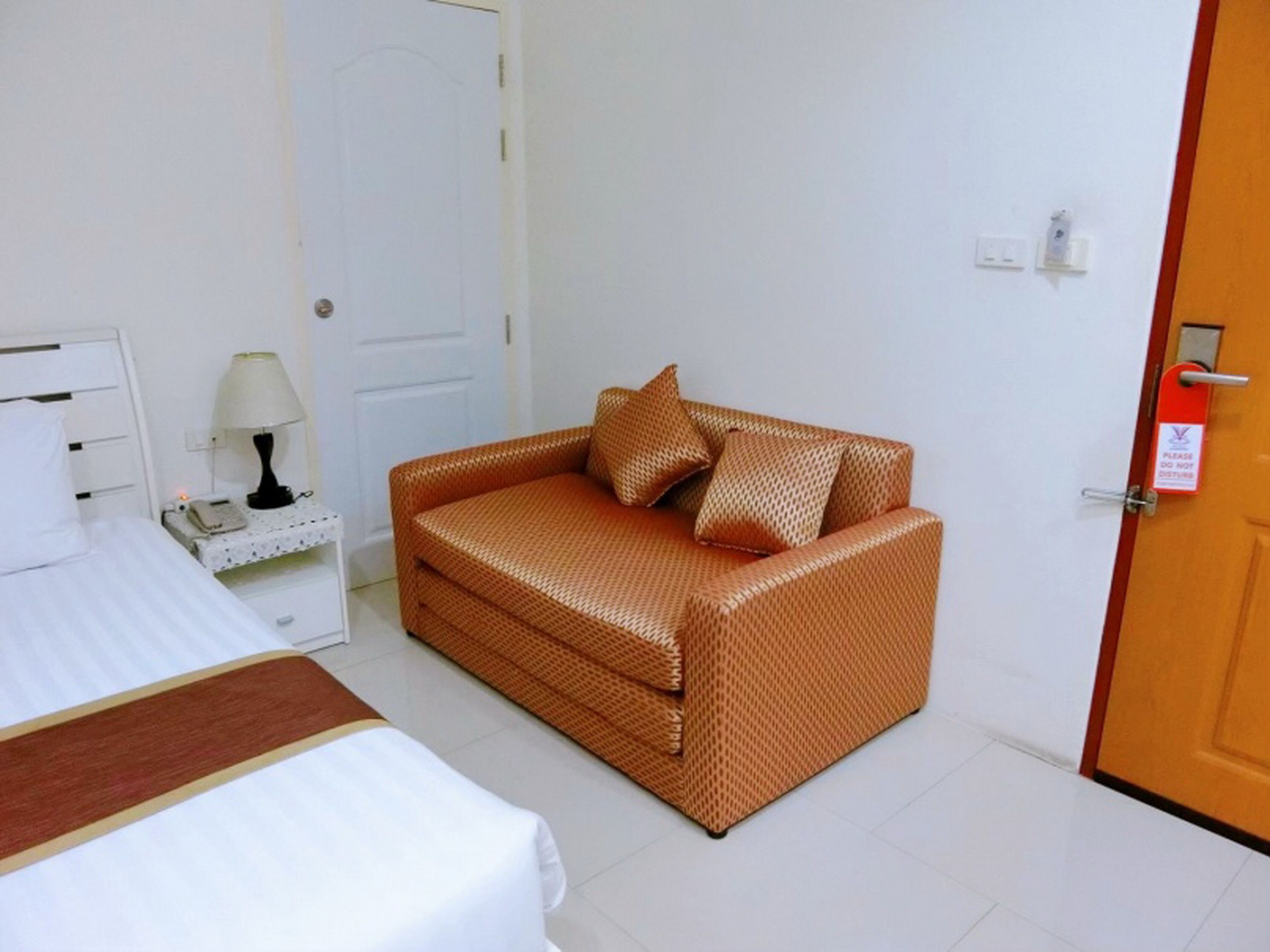 A small, clean room with a white bed, a wicker sofa with cushions, a nightstand with a lamp, and a door with a red sign.