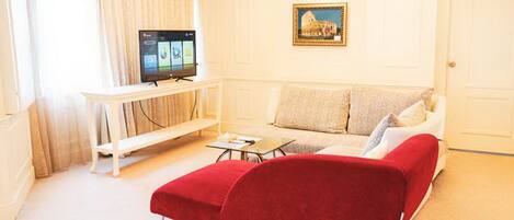 Una sala de estar con un sofá rojo, un sofá blanco, un televisor sobre un mueble bajo y un cuadro en la pared.