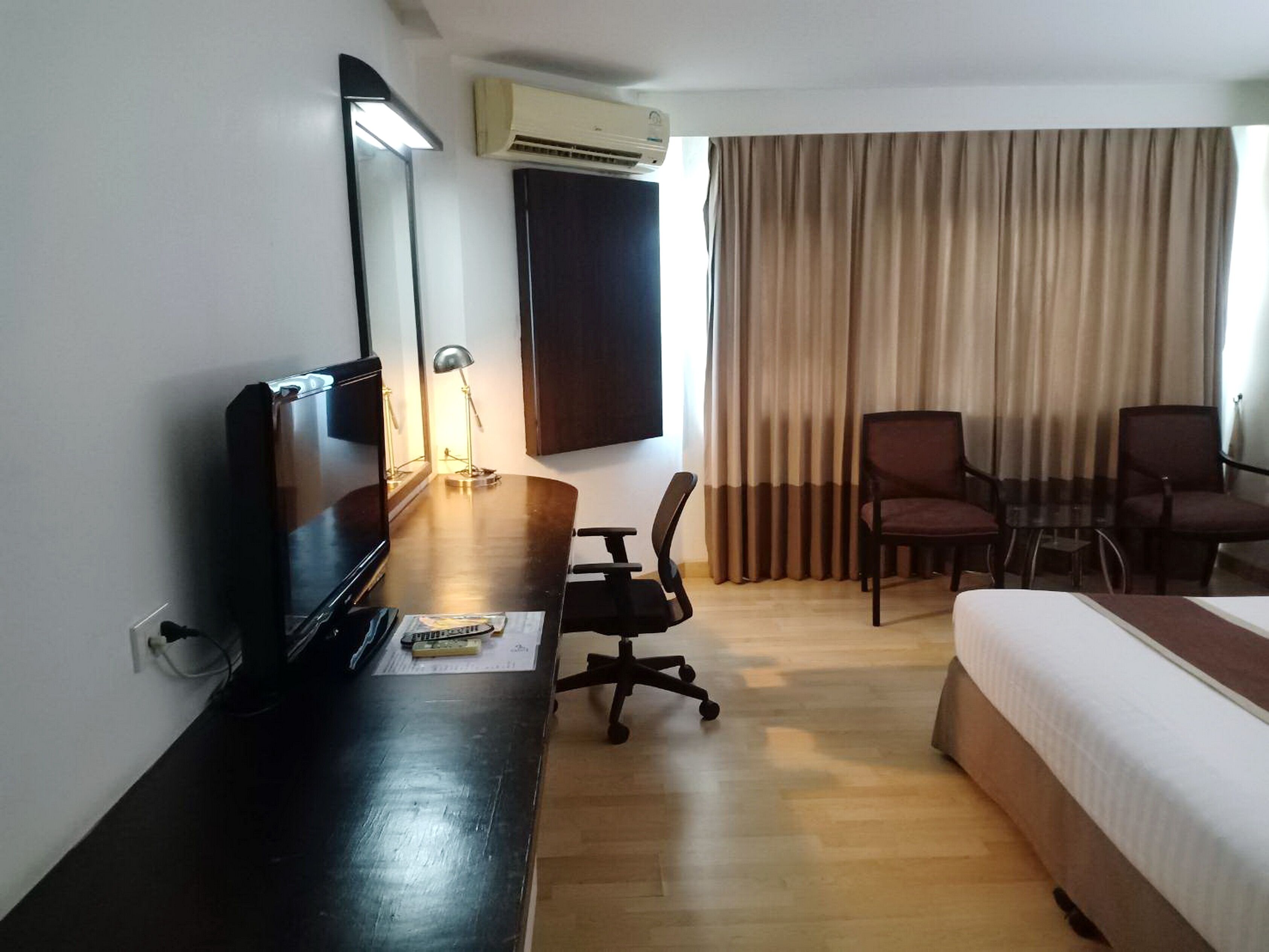 A hotel room with a bed, a desk with a chair, a TV, an air conditioner, and a window with curtains.