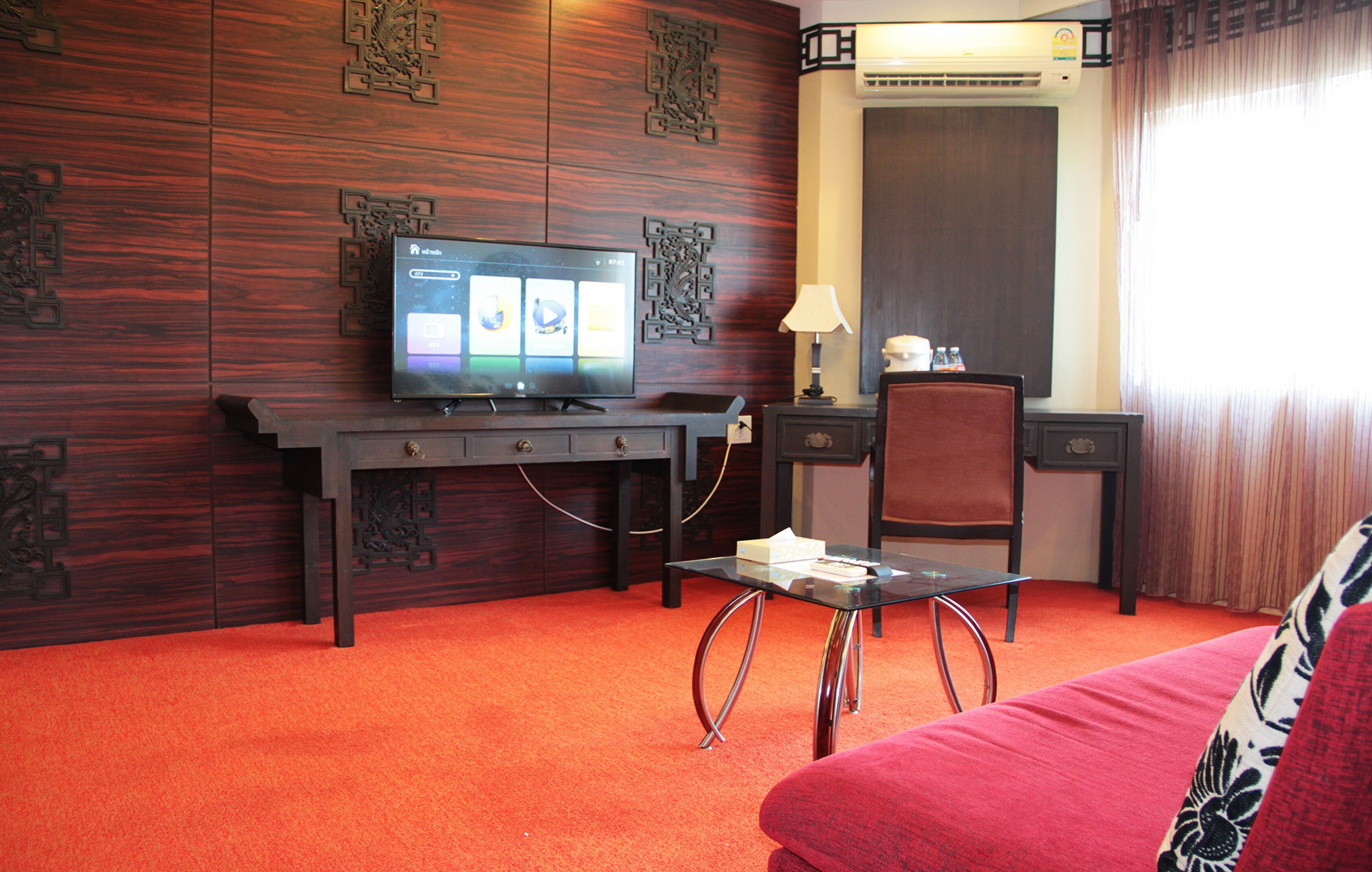 Quarto de hotel com televisão sobre uma escrivaninha de madeira, uma cadeira, uma mesinha e um sofá vermelho.