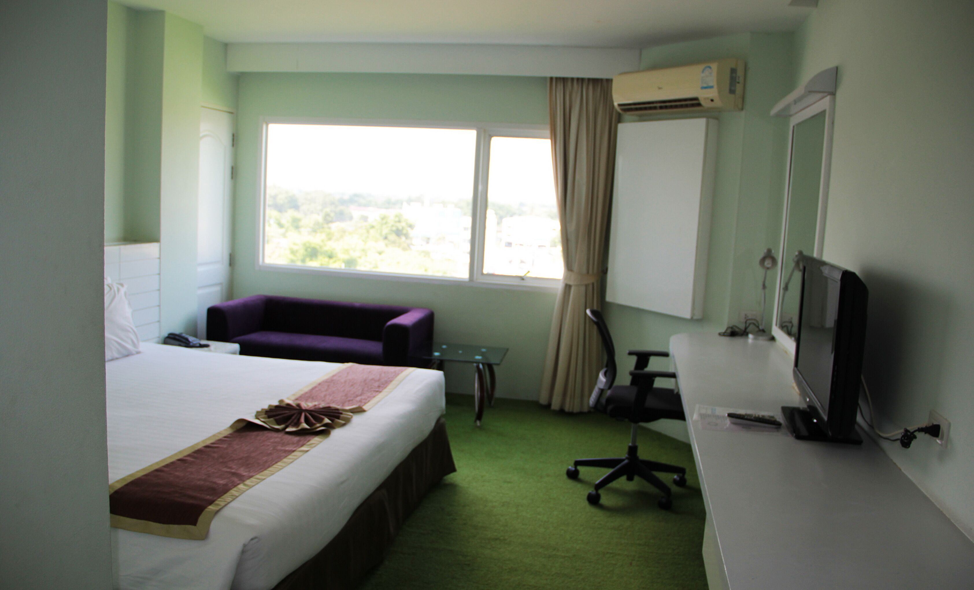 A hotel room with a bed, a purple sofa, a desk with a computer, and a window with a view of greenery.