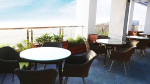 Un espace extérieur aménagé en terrasse, avec des tables rondes et des chaises en osier, entouré de plantes en pot et doté de grandes fenêtres offrant une vue sur le paysage urbain.