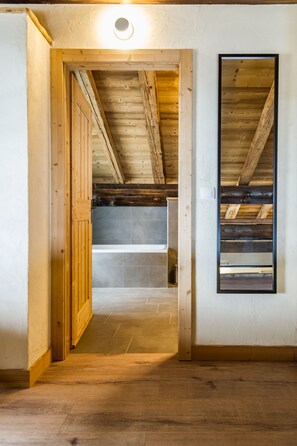 Un corridoio stretto con travi in legno e pavimento piastrellato che conduce a una stanza con soffitto spiovente e una finestra.
