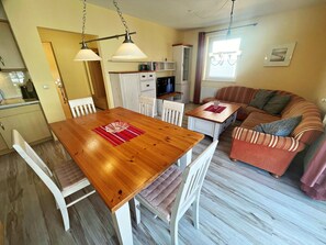 Uma sala de estar acolhedora com uma mesa de jantar de madeira, cadeiras brancas, um sofá listrado e uma cozinha ao fundo.