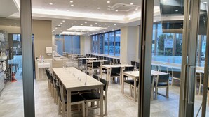 A modern dining area with multiple tables and chairs, a kitchen area with a sink and countertop, and large windows offering a view of the street. - Kuretake Inn Premium Meieki Minami (Nagoya)