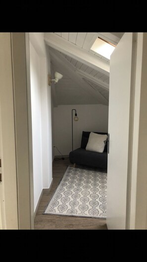 Un pasillo estrecho que da a un dormitorio con techo inclinado, un suelo estampado, una cama con almohadones y una lámpara de pie.