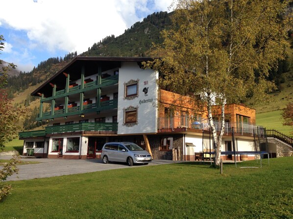 Un immeuble à plusieurs étages doté d’un balcon, avec une voiture garée devant et un paysage montagneux en arrière-plan.