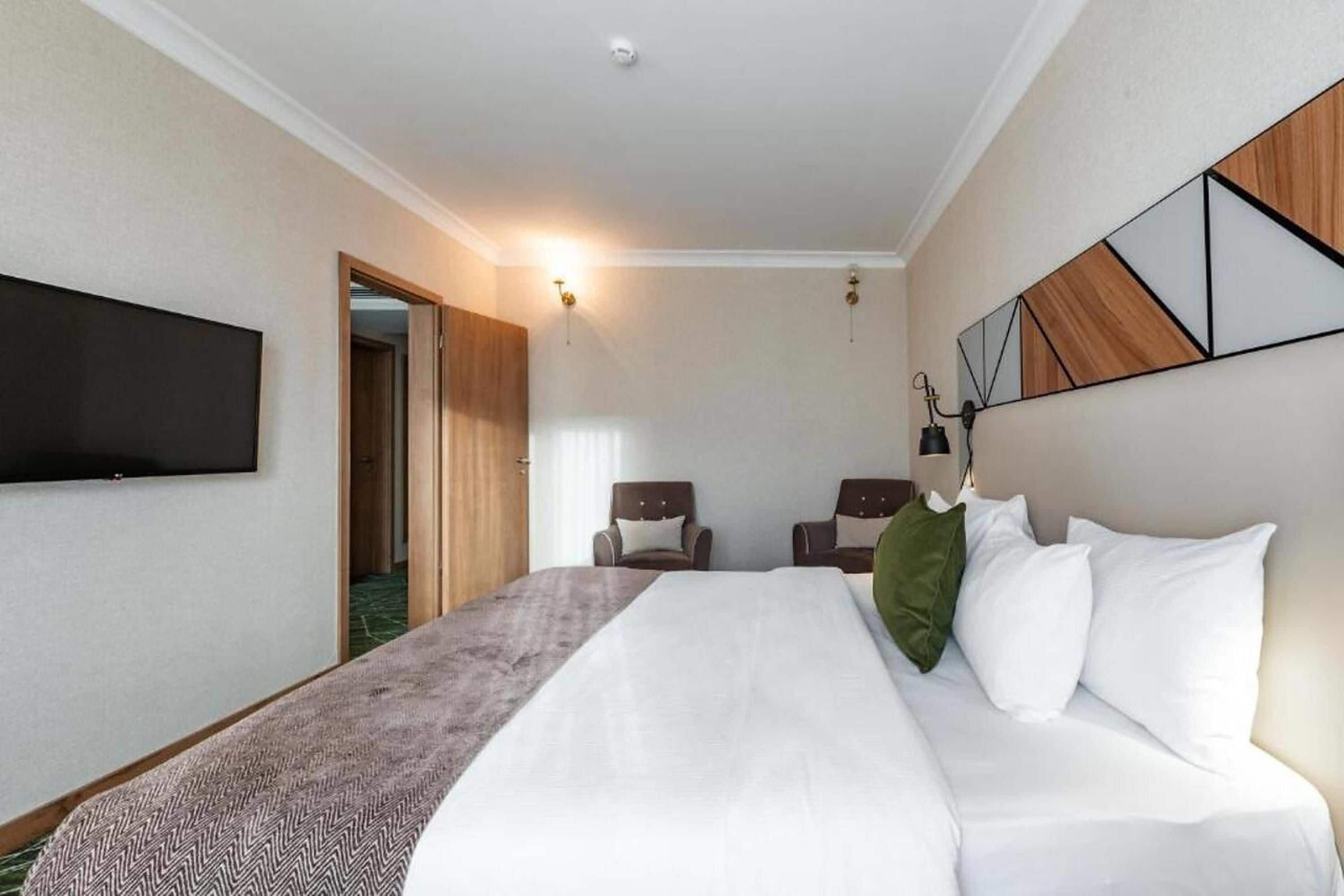 A hotel room with a bed, two chairs, a TV mounted on the wall, and a door leading to another room.