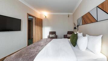 A hotel room with a bed, two chairs, a TV mounted on the wall, and a door leading to another room.