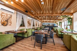 Een restaurant met houten balken in het plafond, groene bekleding op de stoelen en een bar op de achtergrond.