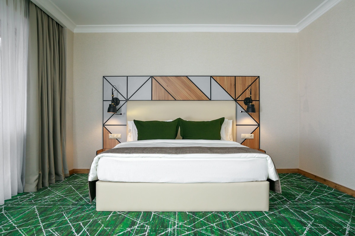Una habitación de hotel con una cama grande, alfombra estampada verde y blanca, un cabecero geométrico y dos lámparas de noche.