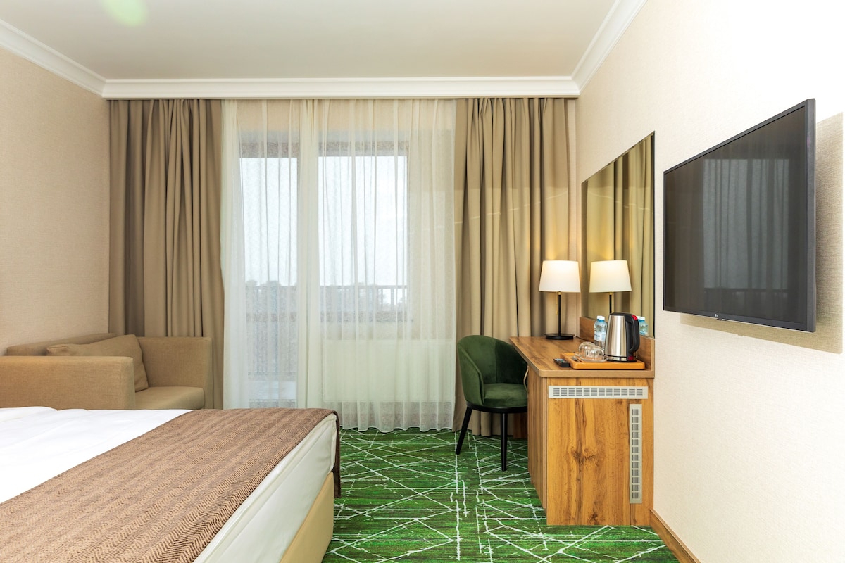 Habitación de hotel con cama, escritorio con lámpara, silla, televisor y ventana con cortinas.