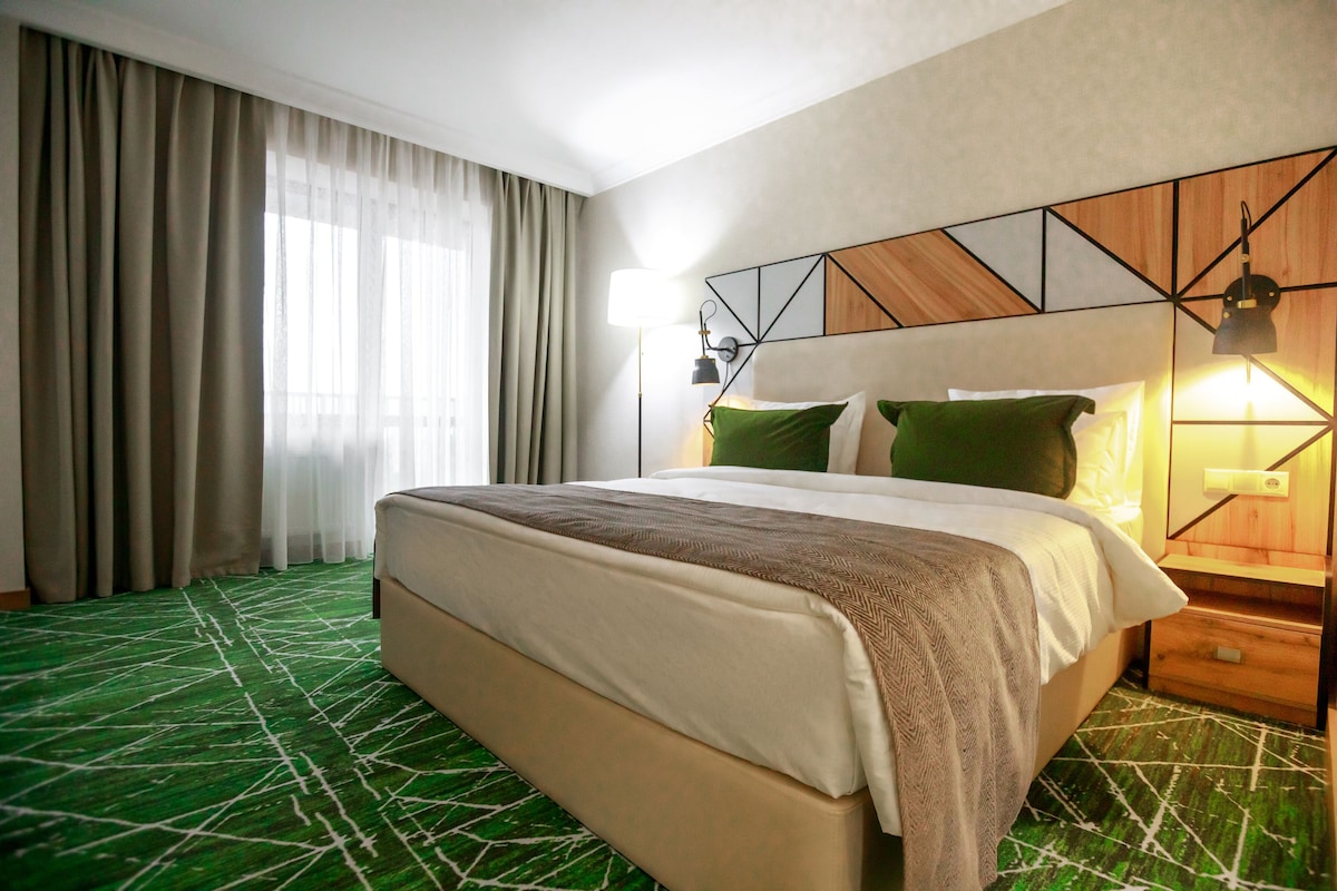 Habitación de hotel con una cama grande, una mesita de noche, una lámpara y una ventana con cortinas.