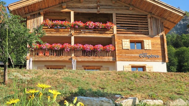 A wooden building with balconies adorned with flowers, a sign reading 'Les Congères,' and a mountainous backdrop.