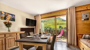 A dining area with a table set for two, a television on the wall, and a scenic view through the glass door.