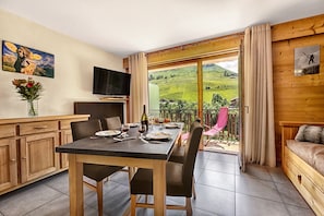 A dining area with a table set for two, a television on the wall, and a scenic view through the glass door.