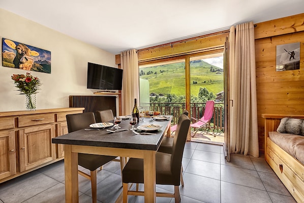 A dining area with a table set for two, a television on the wall, and a scenic view through the glass door. - Beautiful, bright 2-room flat for 4 people (Le Grand Bornand)