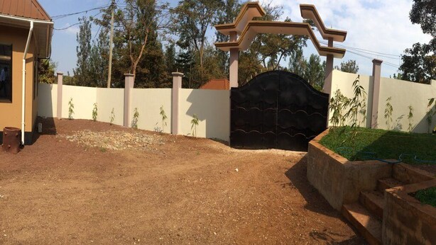 A gated entrance with a decorative archway, a dirt driveway, and a landscaped area with plants. - Sumawe Suites Karatu (Karatu)