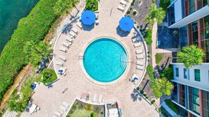 En vy från ovan av en rund pool omgiven av solstolar och parasoller, med en byggnad intill poolen.