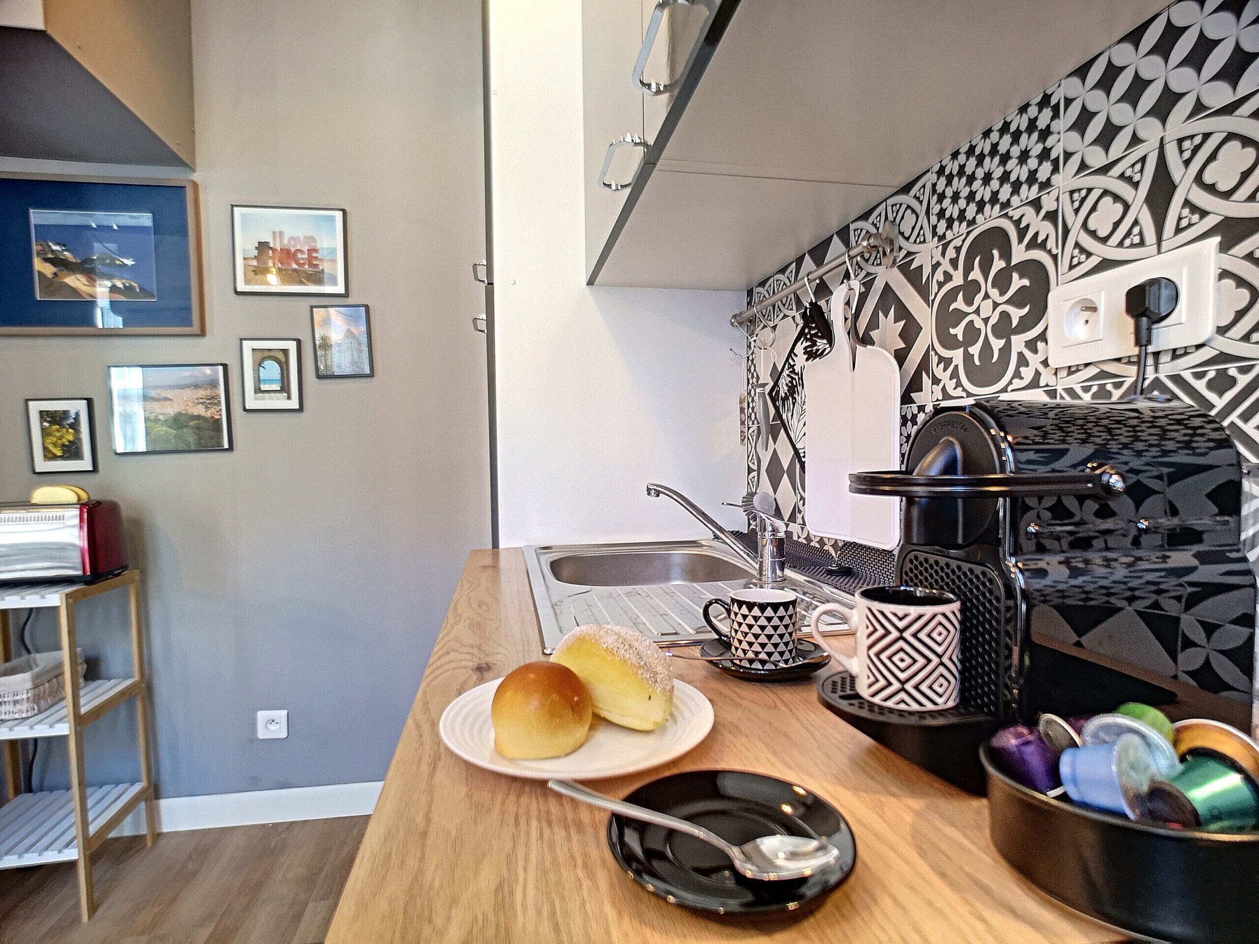 A kitchen with a wooden countertop, a sink, and a patterned backsplash. There are framed pictures on the wall and a shelf with books and a lamp.