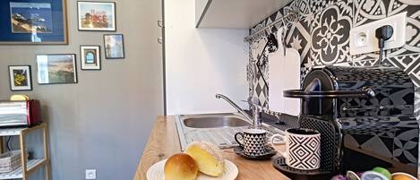 A kitchen with a wooden countertop, a sink, and a patterned backsplash. There are framed pictures on the wall and a shelf with books and a lamp.