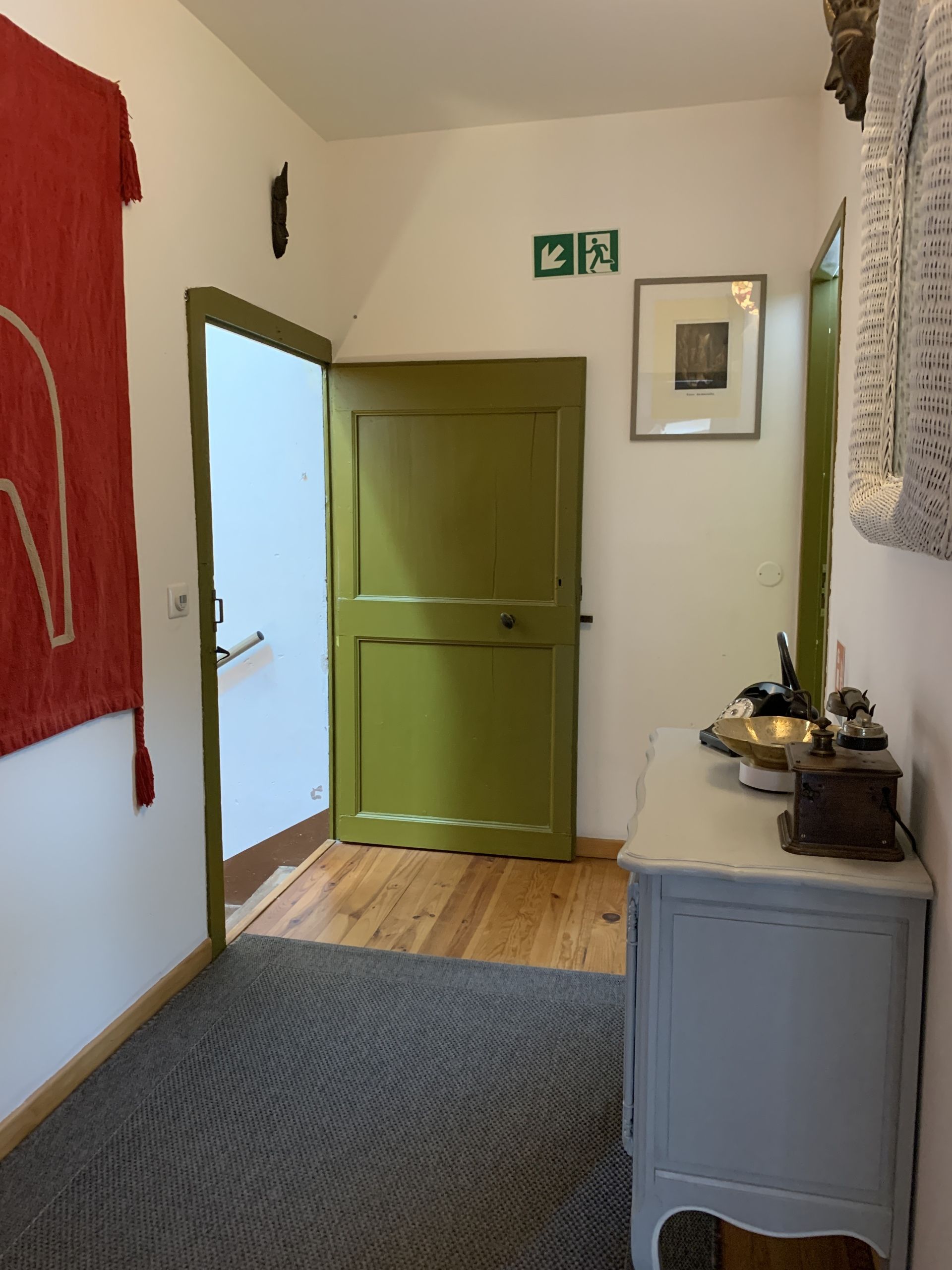 En gang med en grøn dør, et billede i ramme på væggen og et lille bord med en lampe og en skål.