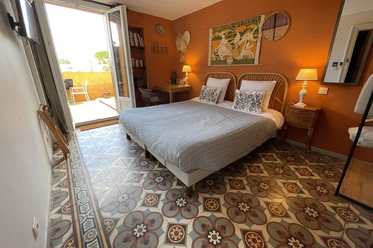 A bedroom with a bed, a chair, a table, and a door leading to an outdoor area.